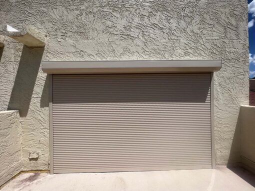 A large, closed beige fire resistant shutter installed against a textured, stucco wall on a sunny day.